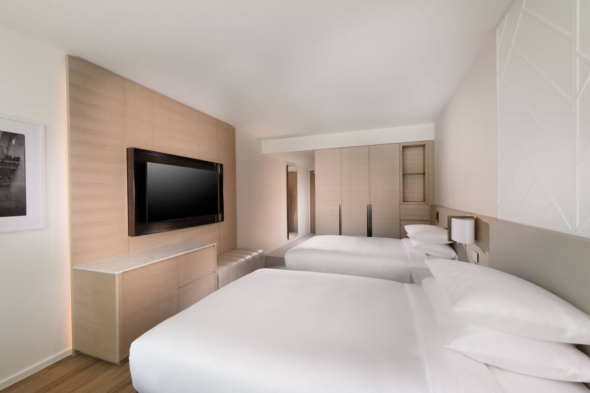 Moderni hotellihuone, jossa on seinälle asennettu taulutelevisio, puinen kaappi ja kaksi sänkyä valkoisilla vuodevaatteilla.