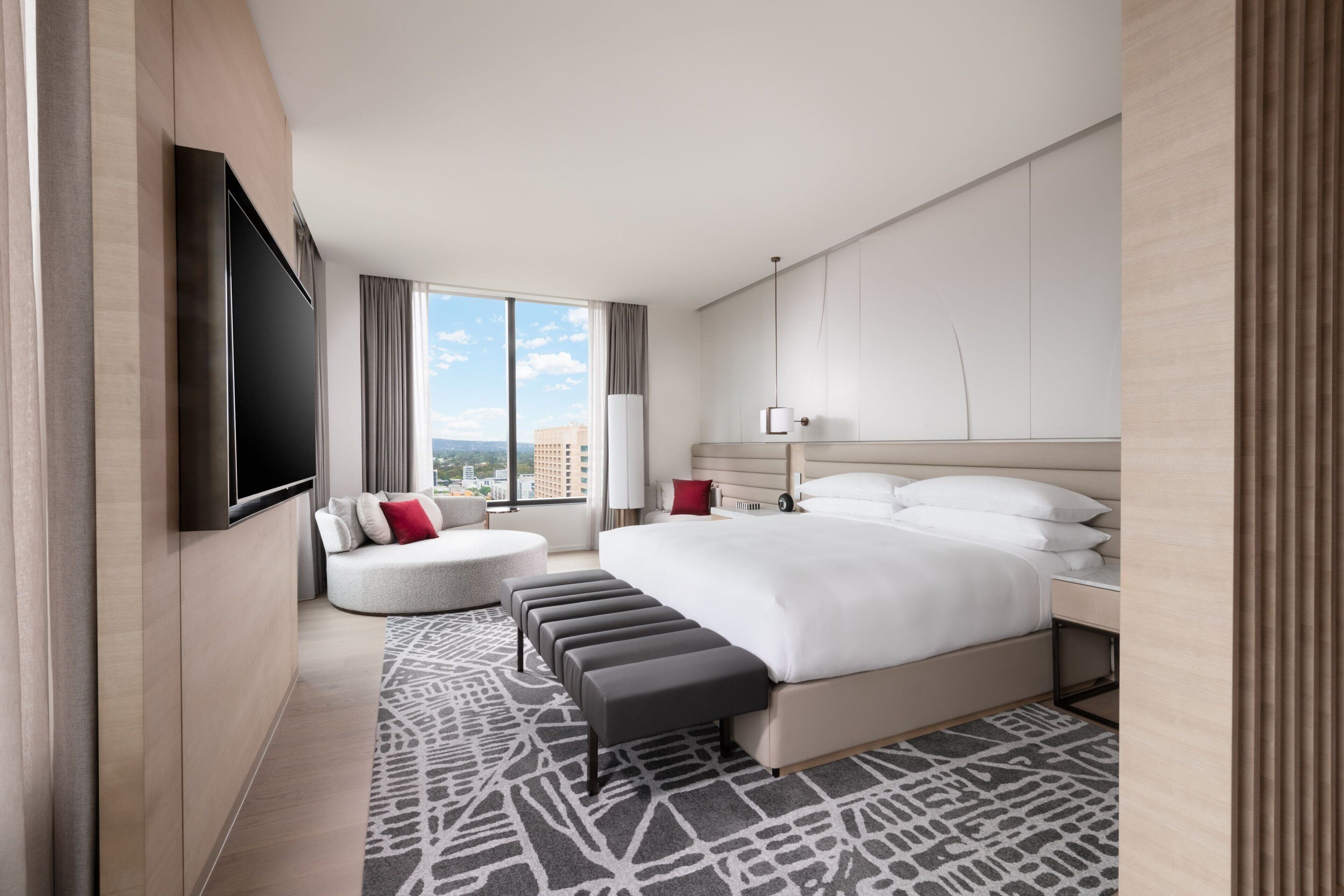 A modern hotel room with a large bed, a flat-screen TV mounted on the wall, a seating area with a sofa and ottoman, and a large window offering a city view.