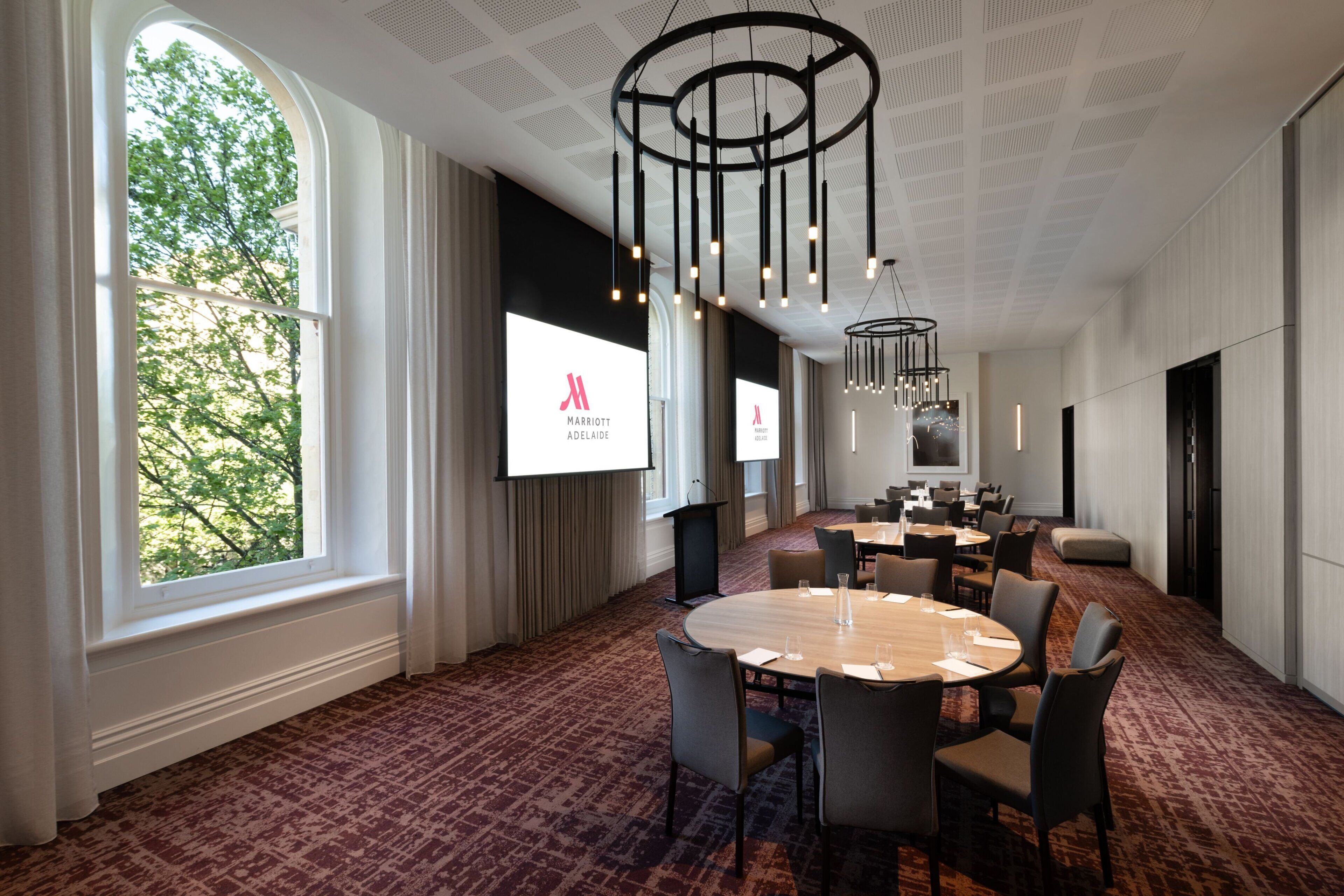 A conference room with a large round table, multiple chairs, a projector screen displaying the Marriott logo, and a large window with a view of trees.