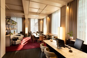 A modern hotel room with a large dining table, comfortable seating, a long window with curtains, and a bookshelf with decorative items.