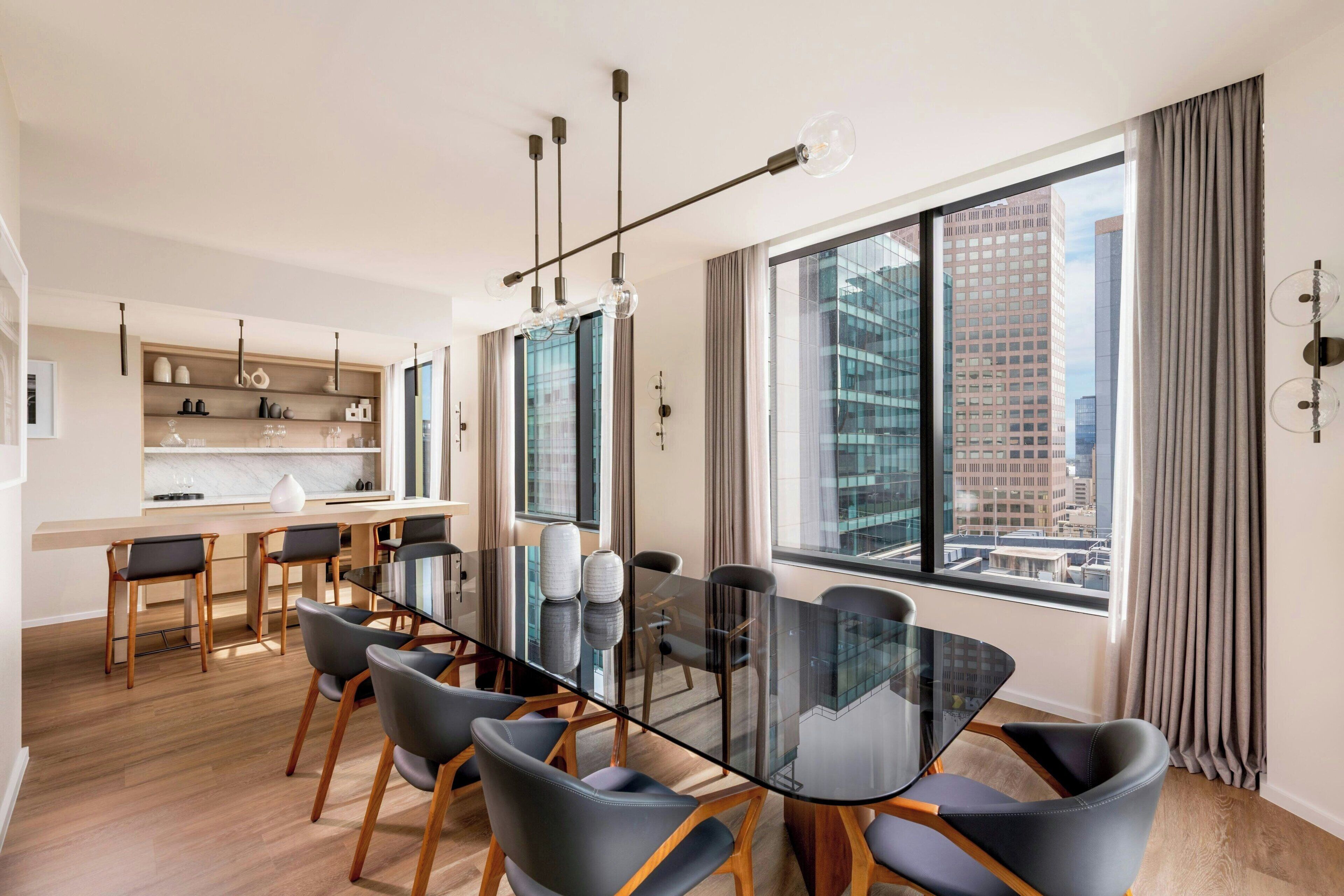 A modern dining area with a glass table and chairs, a kitchen with wooden cabinets, and large windows offering a city view.
