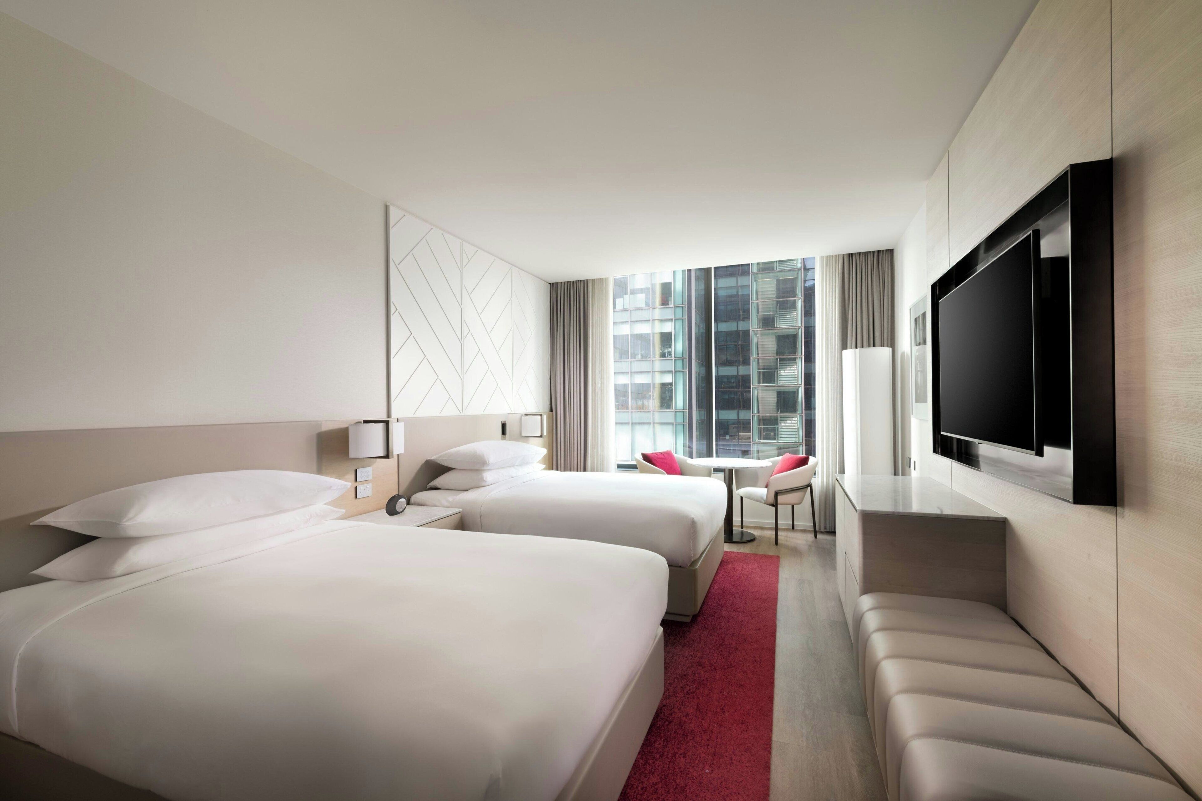 Ein Hotelzimmer mit zwei Betten, einem Fernseher, einem Schreibtisch und Blick auf die Stadt.