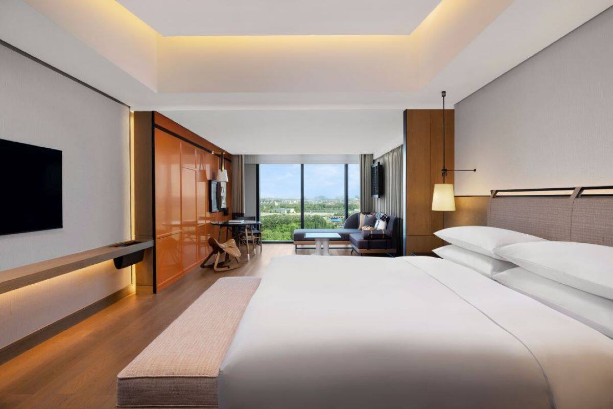 A modern hotel room with a large bed, a flat-screen TV, a seating area with a view, and a large window.