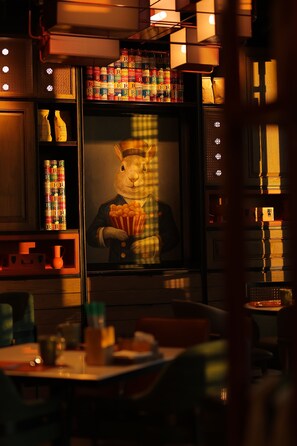 Interior de restaurante con un cuadro grande de un conejo sosteniendo palomitas, estantes con conservas y una mesa con sillas.