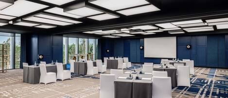 Une salle de conférence équipée de tables et de chaises, d’un écran de projection et de grandes fenêtres.