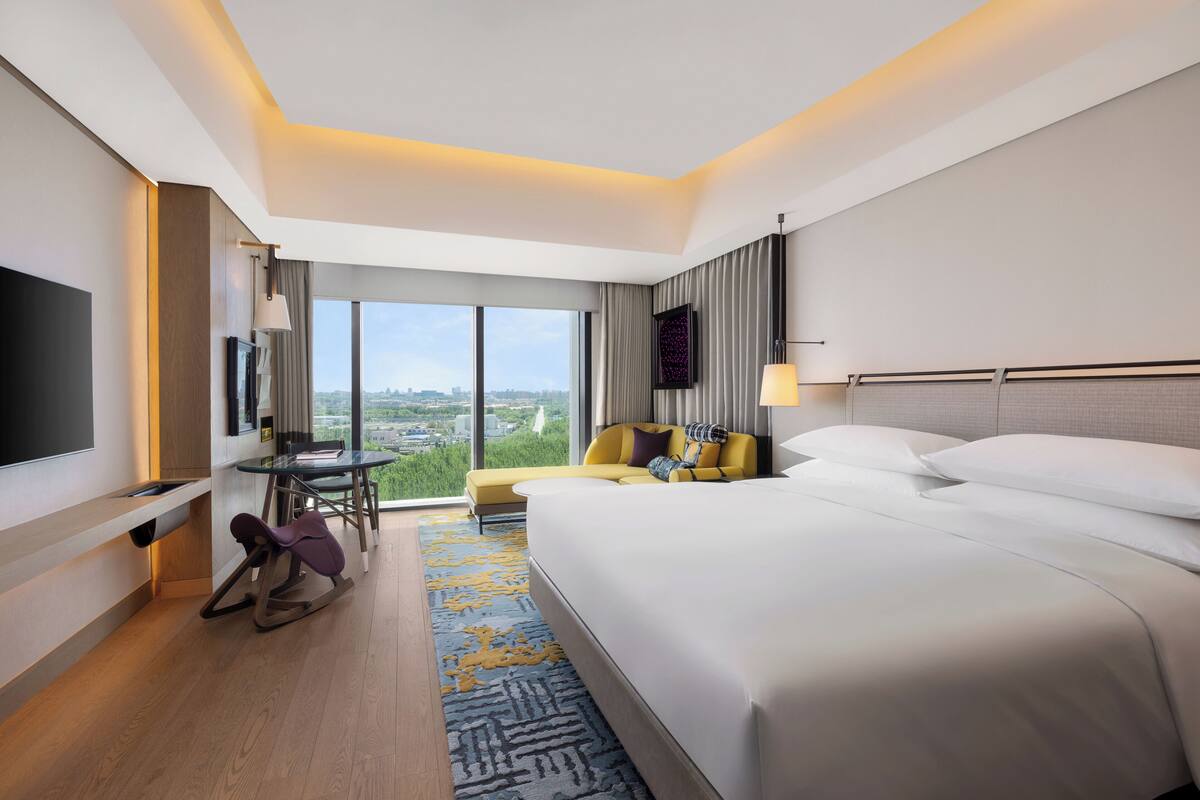 Une chambre d’hôtel moderne dotée d’un grand lit, d’un téléviseur à écran plat, d’un coin repas avec une table et des chaises, et offrant une vue sur la ville grâce à de grandes fenêtres.
