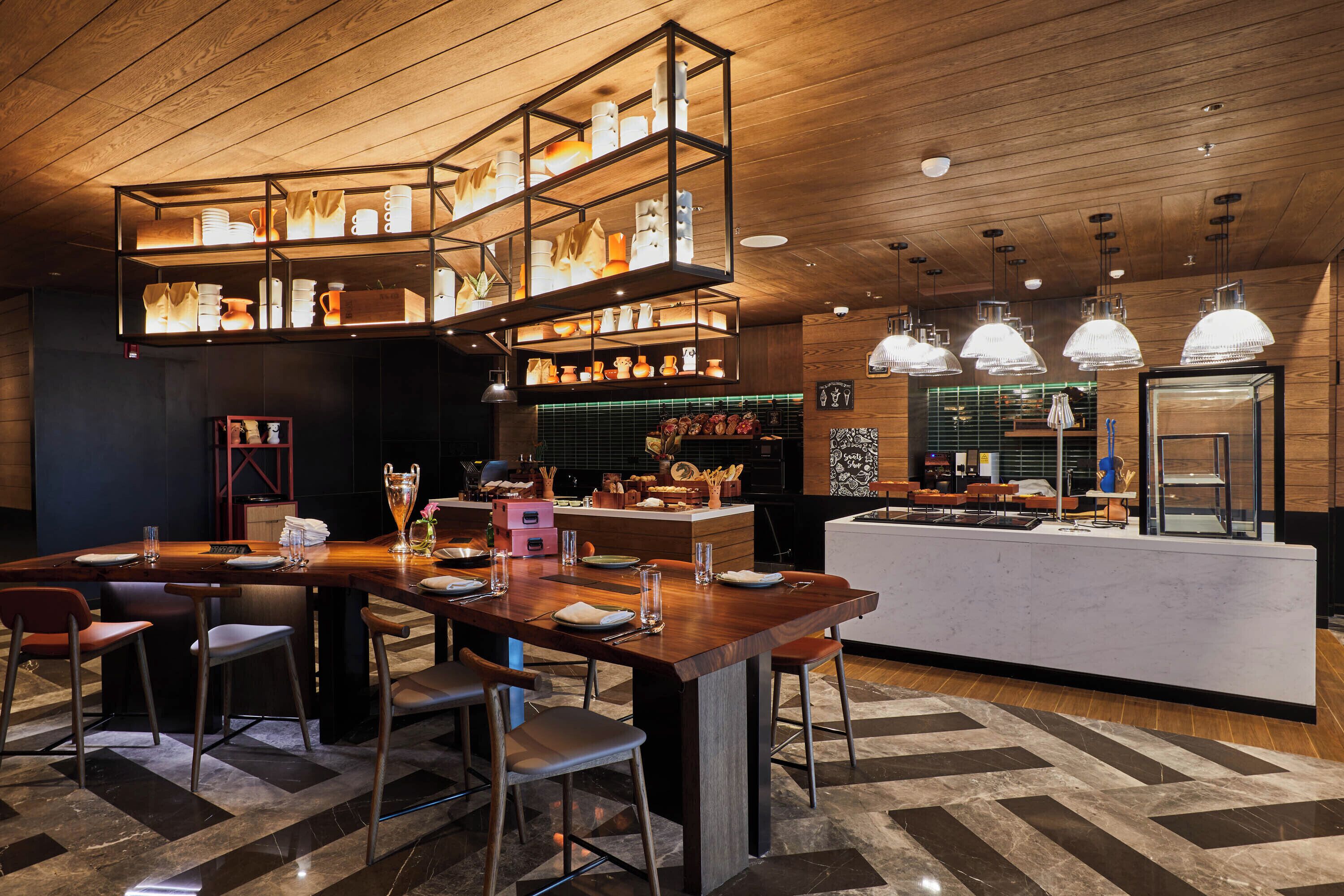 A modern bar area with wooden furniture, a central bar counter, and overhead shelves displaying glasses and bottles.