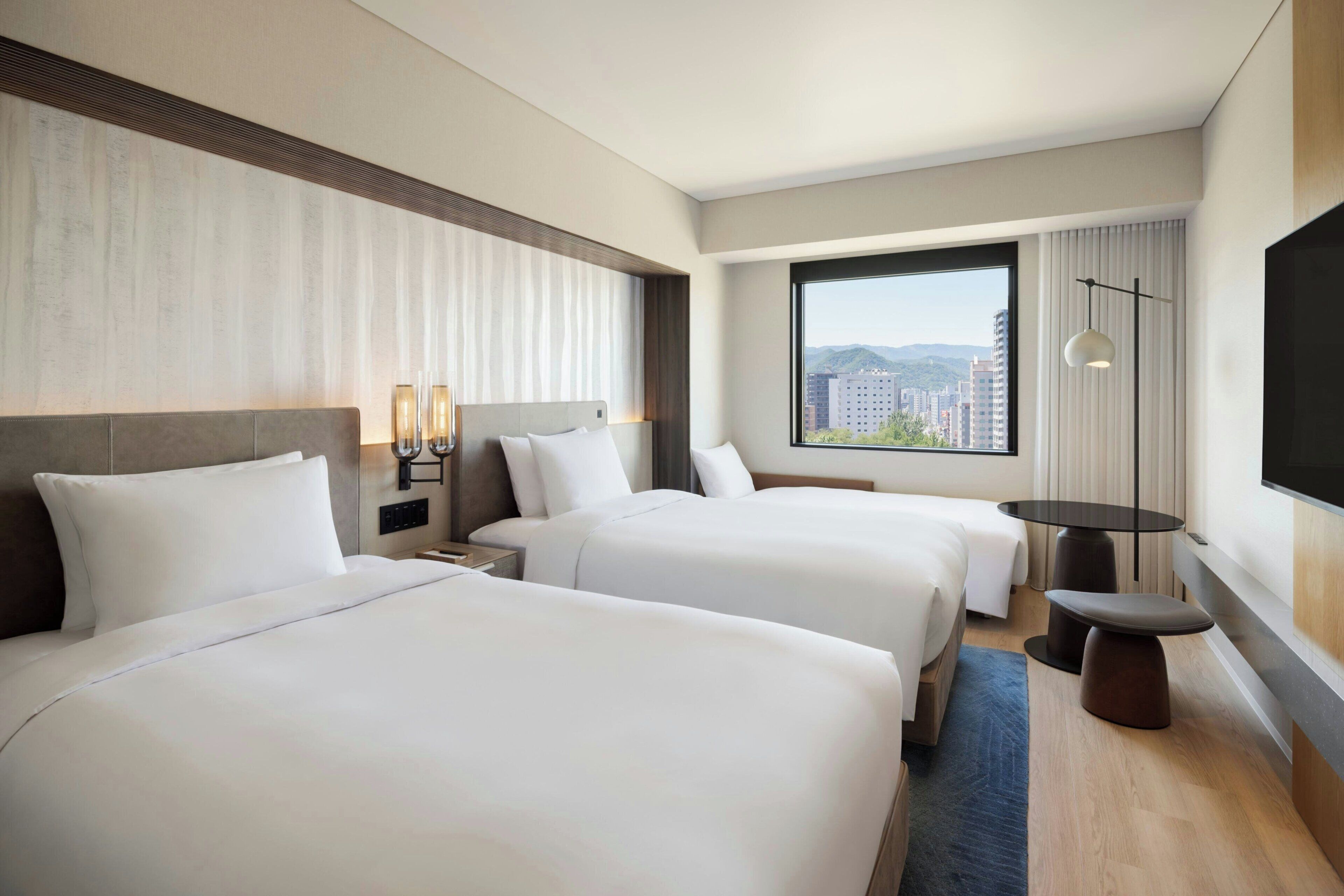 Habitació d'hotel amb dos llits, una finestra gran amb vista a la ciutat, una taula petita i una làmpada penjada a la paret.