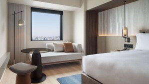 Una habitación de hotel moderna con una cama grande, una zona de estar con sofá y una mesita, un ventanal con vistas a la ciudad y un teléfono en la pared.