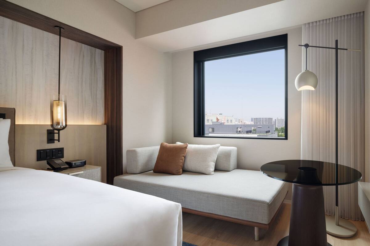 A modern hotel room with a large window, a sofa bed, a bedside table with a lamp, and a small round table.