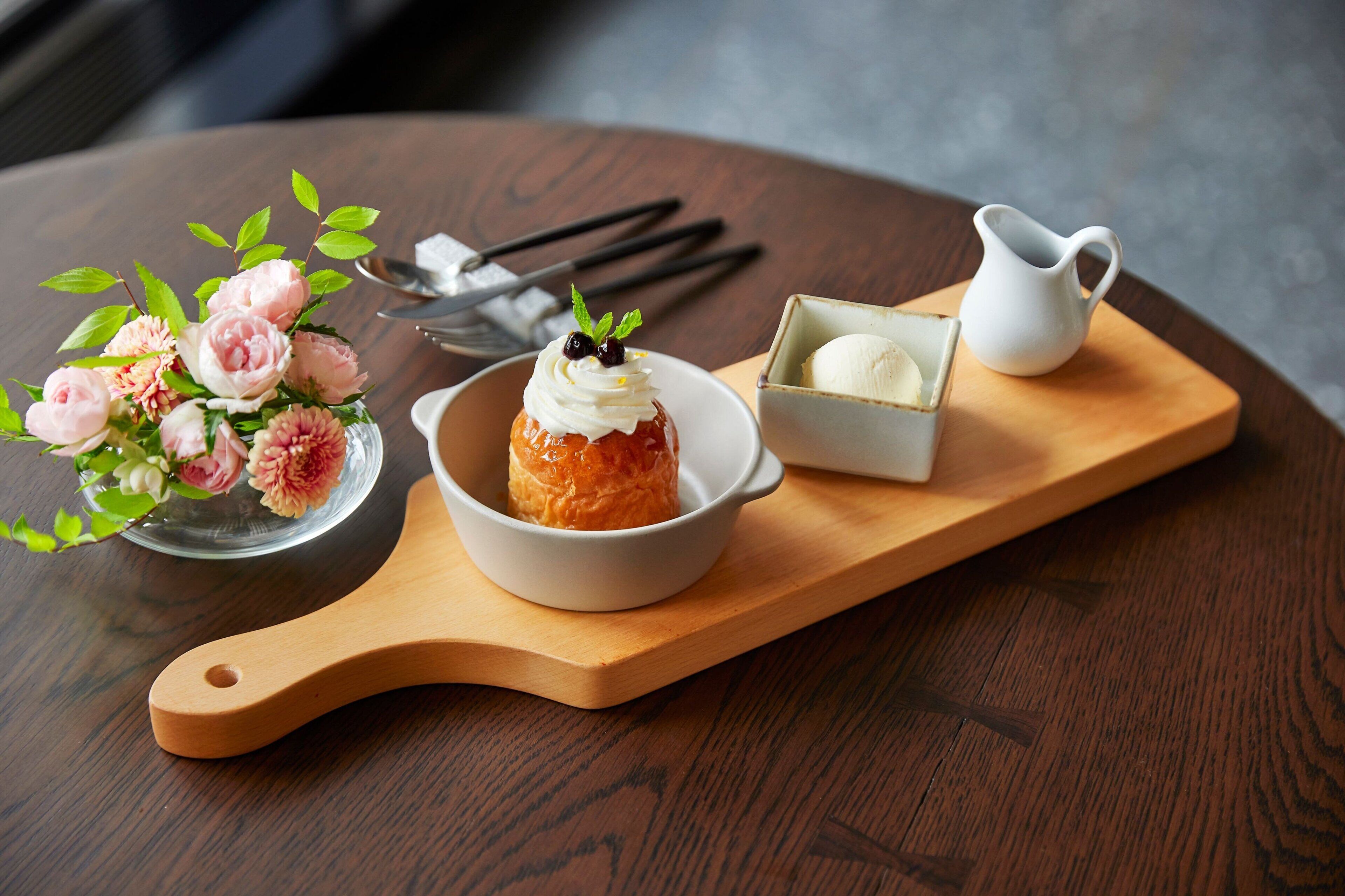 A dessert with whipped cream and a mint leaf on a wooden board, accompanied by a small bowl of ice cream and a pitcher of cream.