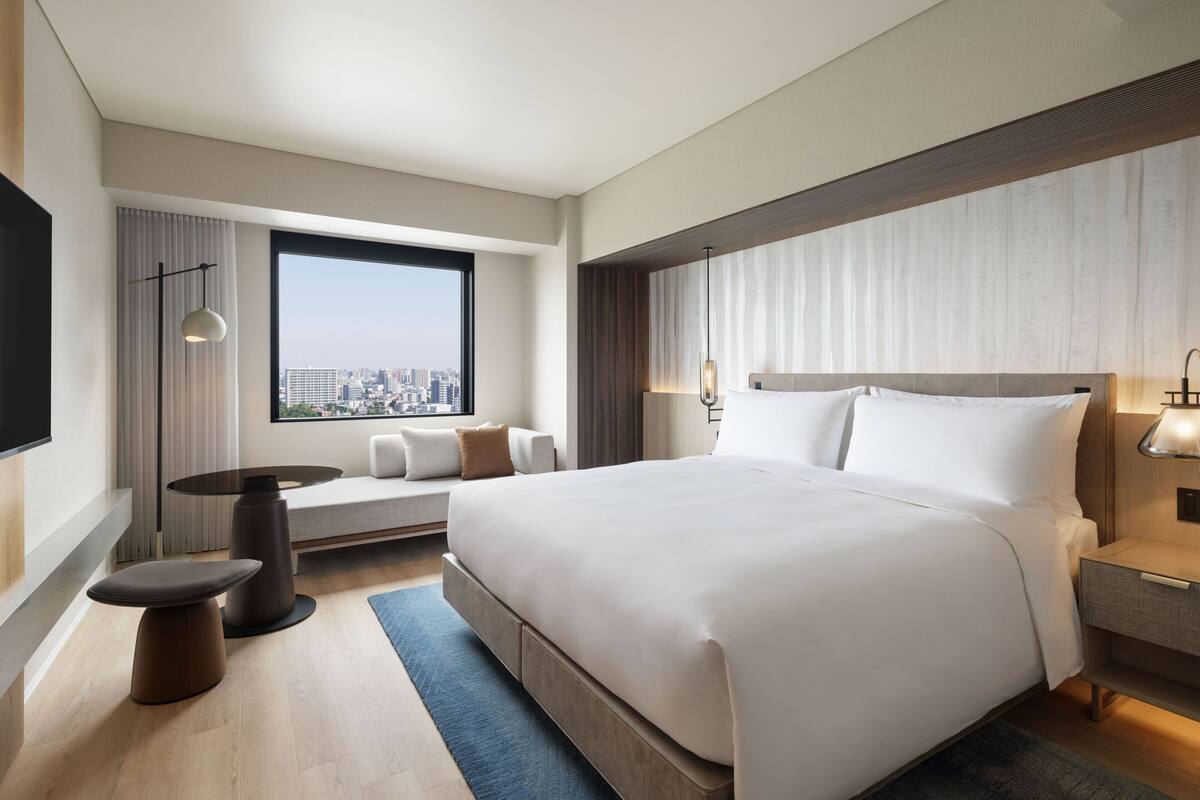 A modern hotel room with a large bed, a seating area with a sofa and armchairs, a small table, and a view of the city through a large window.