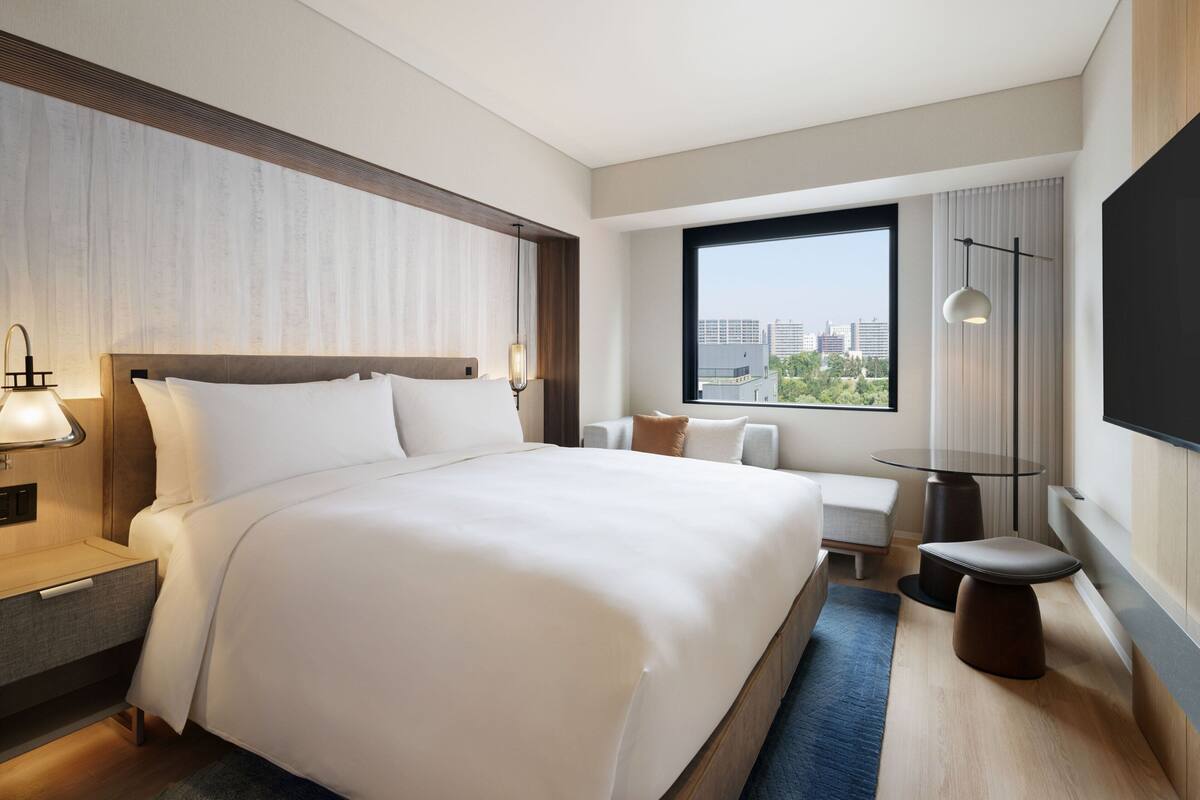 A modern hotel room with a large bed, bedside tables, a desk, and a large window offering a city view.