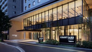 Una entrada de hotel moderna con fachada de vidrio, un cartel que indica 'Courtyard by Marriott Sapporo', y un camino que lleva a la entrada.
