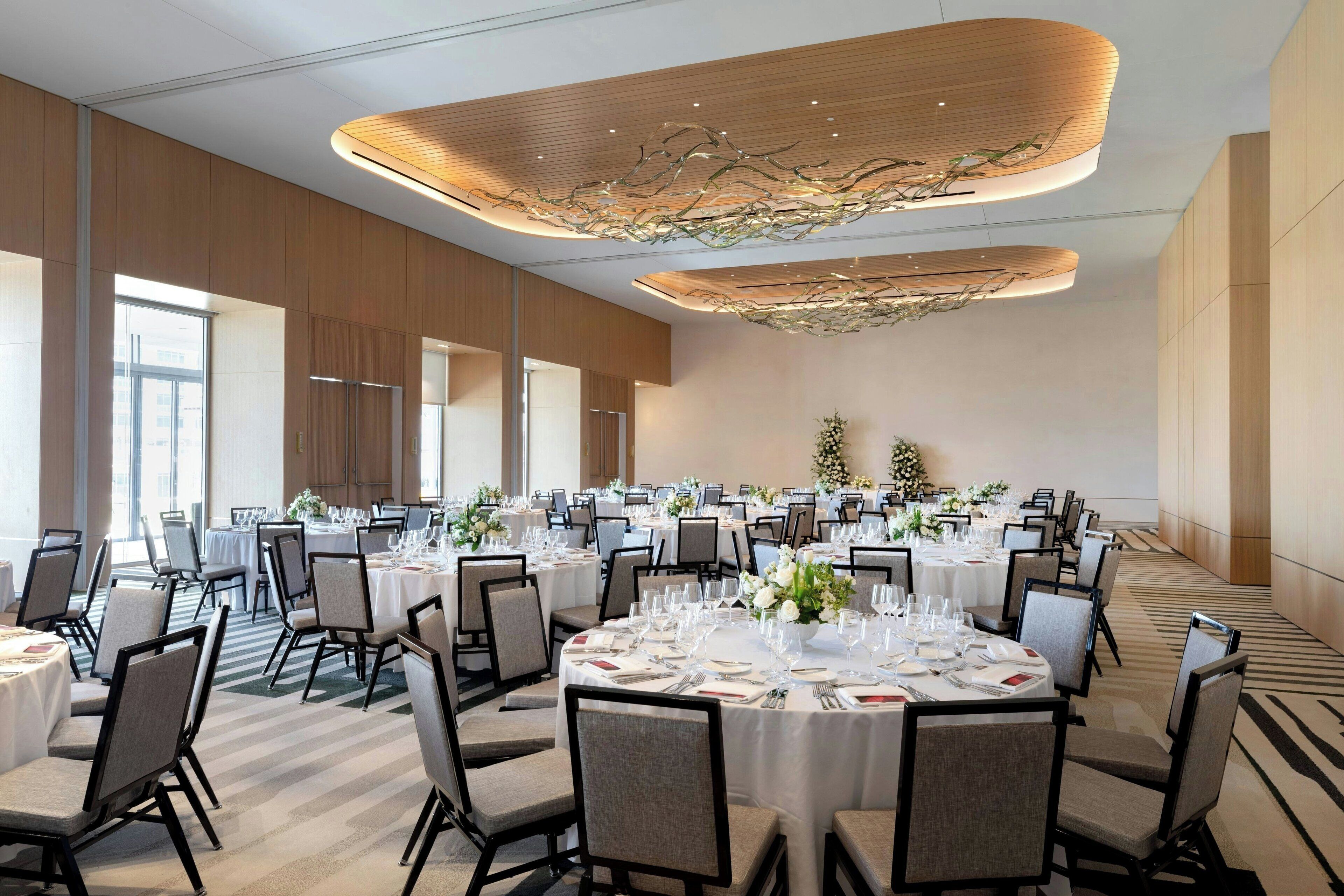 A spacious banquet hall with round tables set for dining, large windows, and a modern ceiling design.