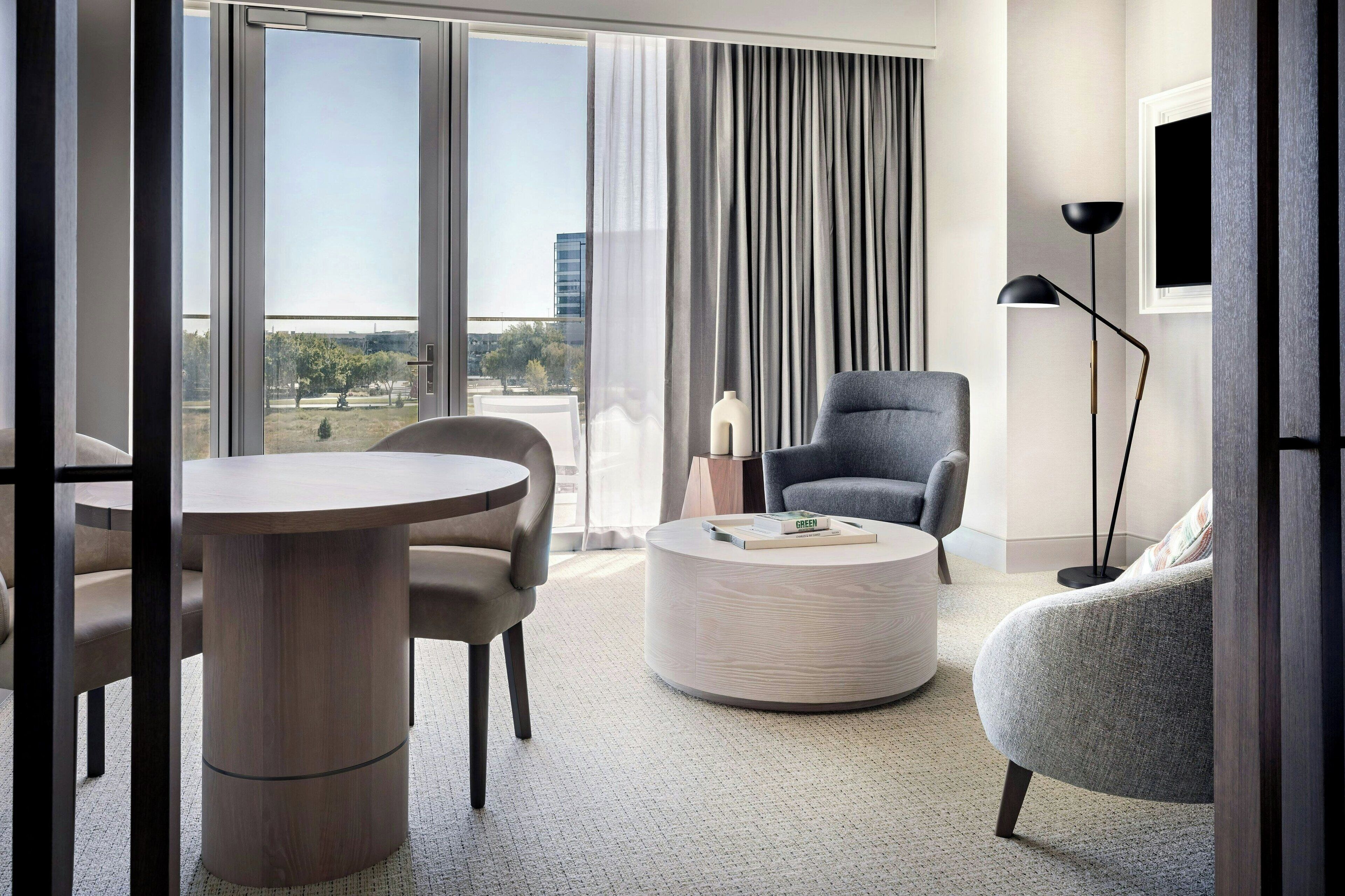 A modern hotel room with a large window, a round coffee table, a grey armchair, and a dining table with chairs.