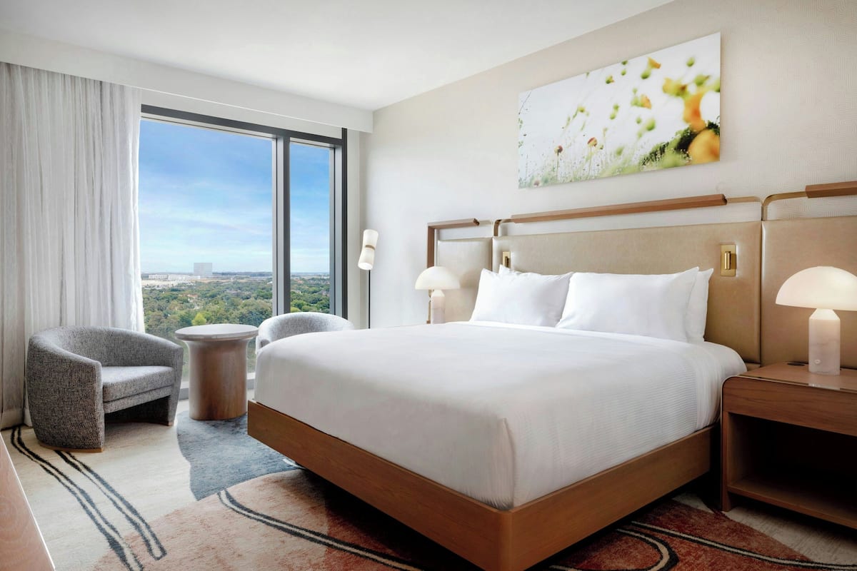 A hotel room with a large bed, a chair, a round table, and a view of the city through large windows.