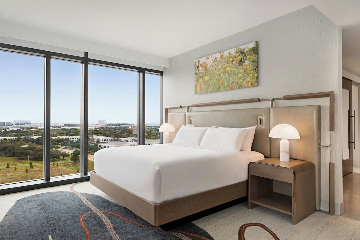 A modern hotel room with a large bed, a nightstand with a lamp, and a city view through large windows.