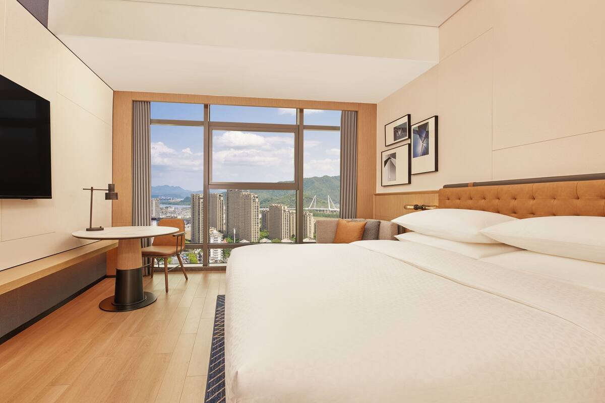 Una habitación de hotel moderna con una cama grande, un escritorio con una silla, un televisor y un amplio ventanal con vista a la ciudad.