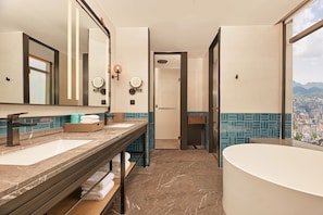 Un baño moderno con un espejo grande, dos lavamanos, una bañera y vistas a un paisaje urbano a través de un ventanal amplio.