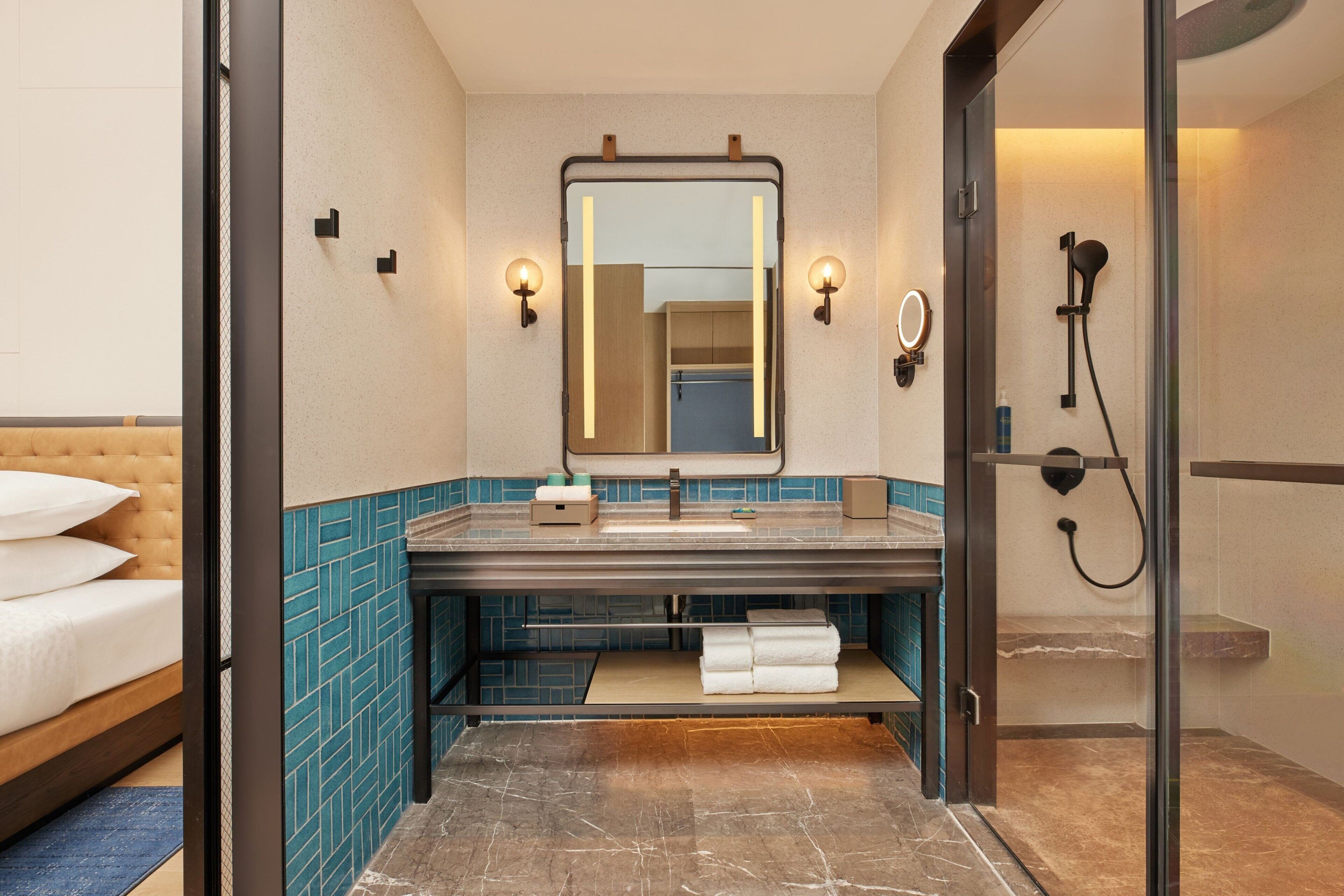 A modern bathroom with a large mirror, a sink, and a shower. There is a bed visible to the left.