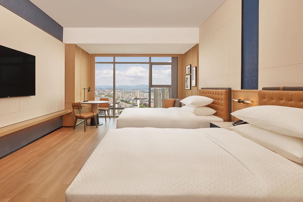 Habitación de hotel con dos camas, un escritorio y un ventanal grande con vistas a la ciudad.
