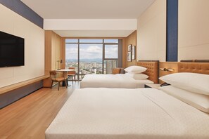 Habitación de hotel con dos camas, un escritorio y un ventanal grande con vistas a la ciudad.