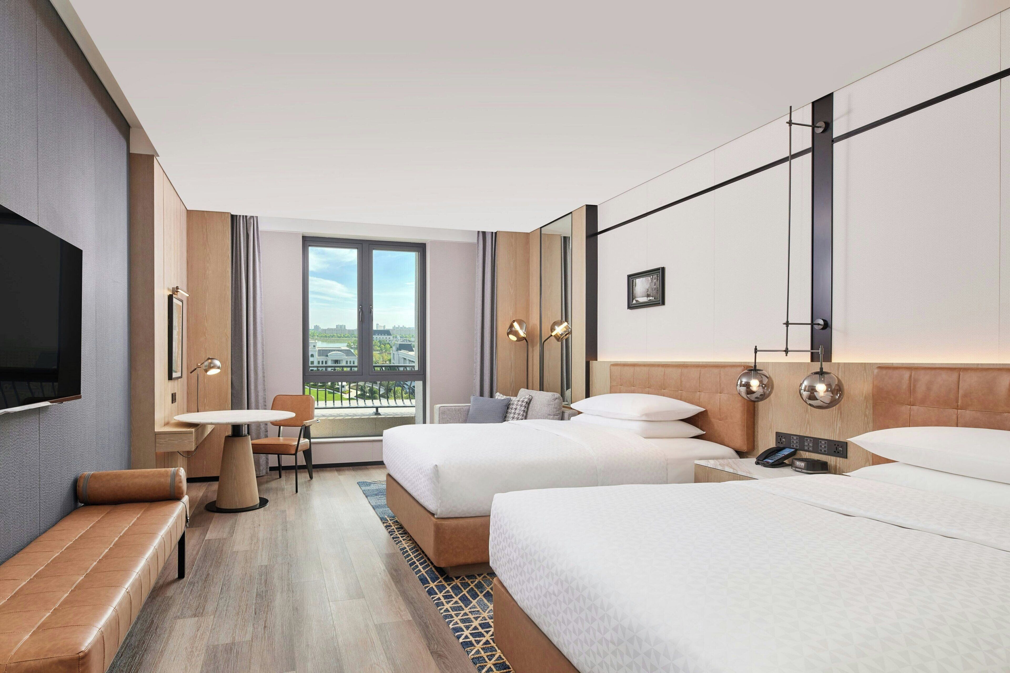 Habitación de hotel con dos camas, un banco, una mesa, un televisor y un ventanal con vistas a la ciudad.