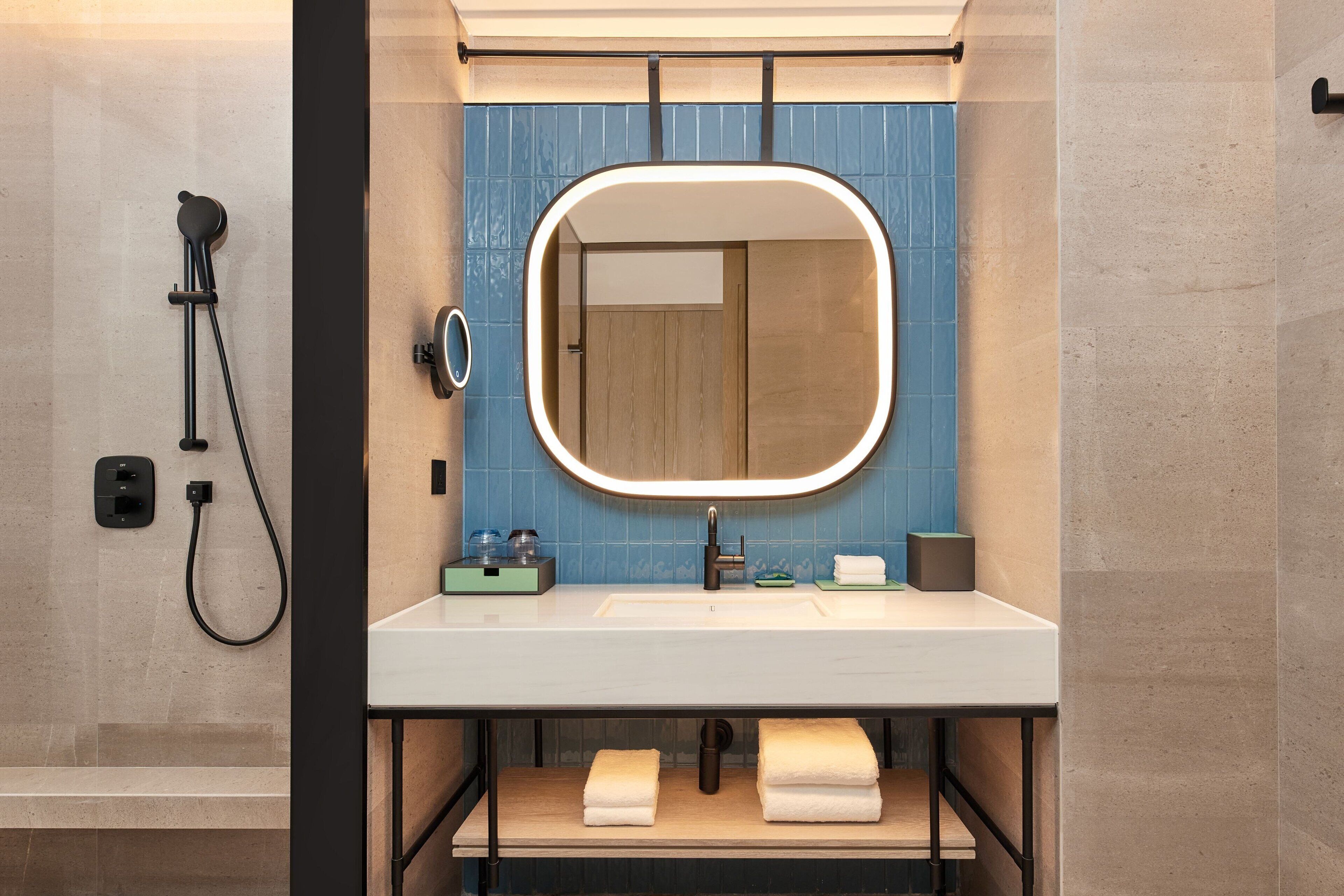 A modern bathroom with a white sink, a mirror with a lighted frame, a handheld showerhead, and a towel rack.