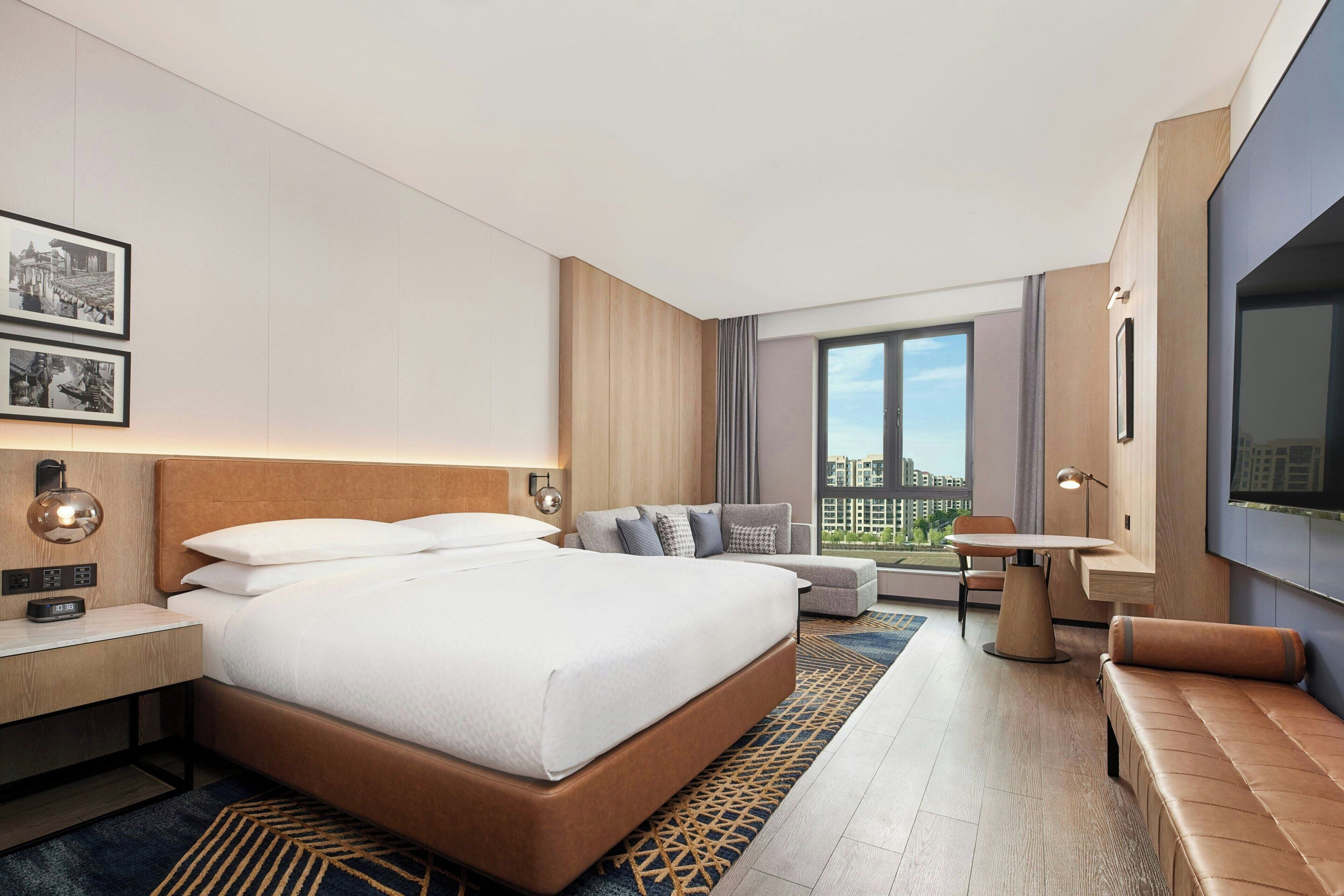 A modern hotel room with a large bed, a sofa, a dining table, and a TV.