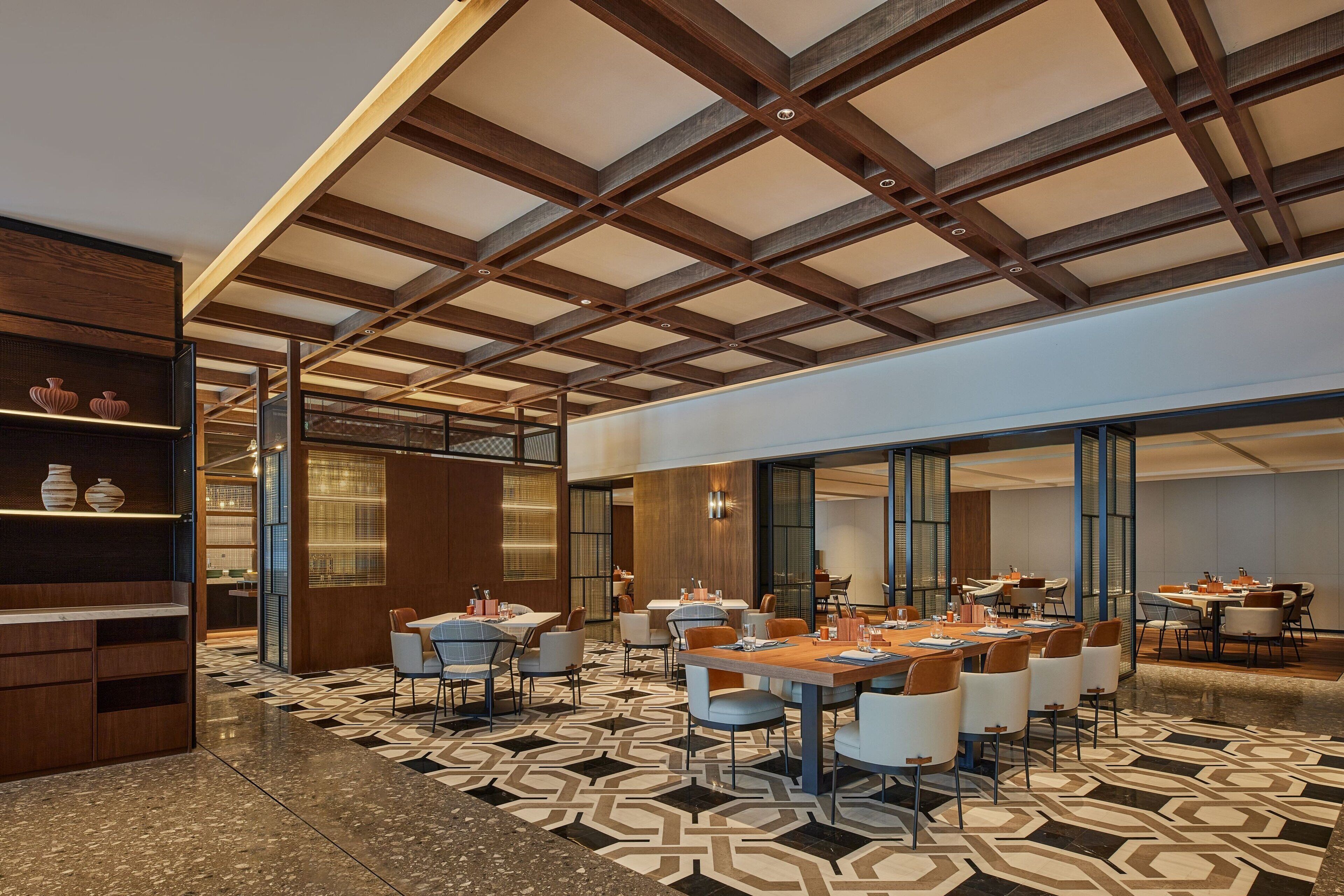 A modern hotel lobby with a high ceiling, geometric patterned floor, and multiple dining areas with tables and chairs.