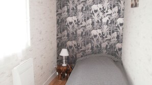 Una habitación pequeña y estrecha con techo inclinado, una cama con una manta gris, una mesita de noche con lámpara y un papel tapiz con motivos de jungla.