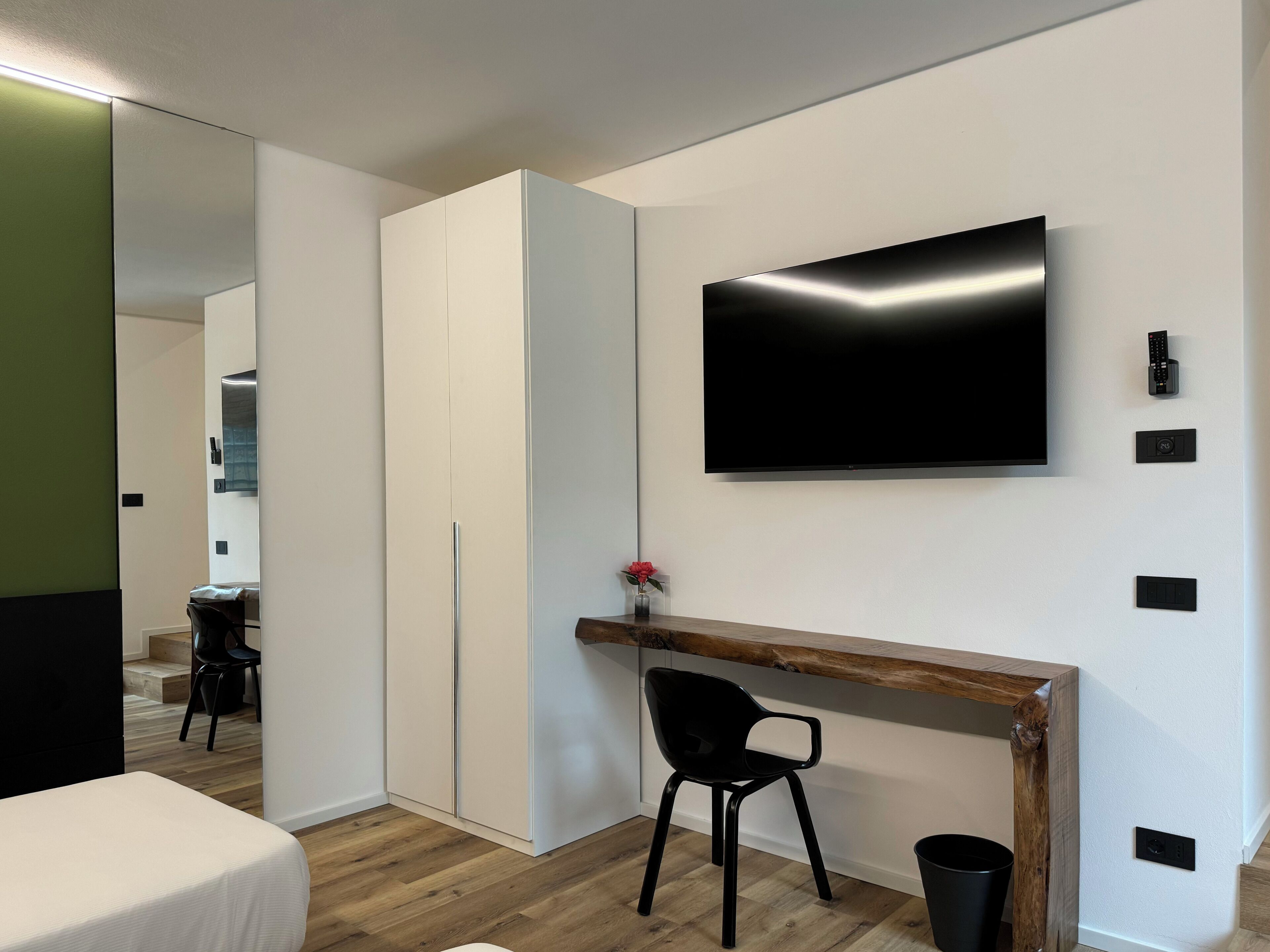A modern hotel room with a large flat-screen TV mounted on the wall, a wooden console table, a black chair, and a small trash bin.