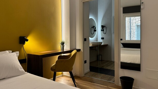 Ein Hotelzimmer mit einem Bett, einem Schreibtisch mit Stuhl, einer kleinen Pflanze, einem Badezimmer mit Waschbecken und Spiegel sowie einem Fenster mit Aussicht.