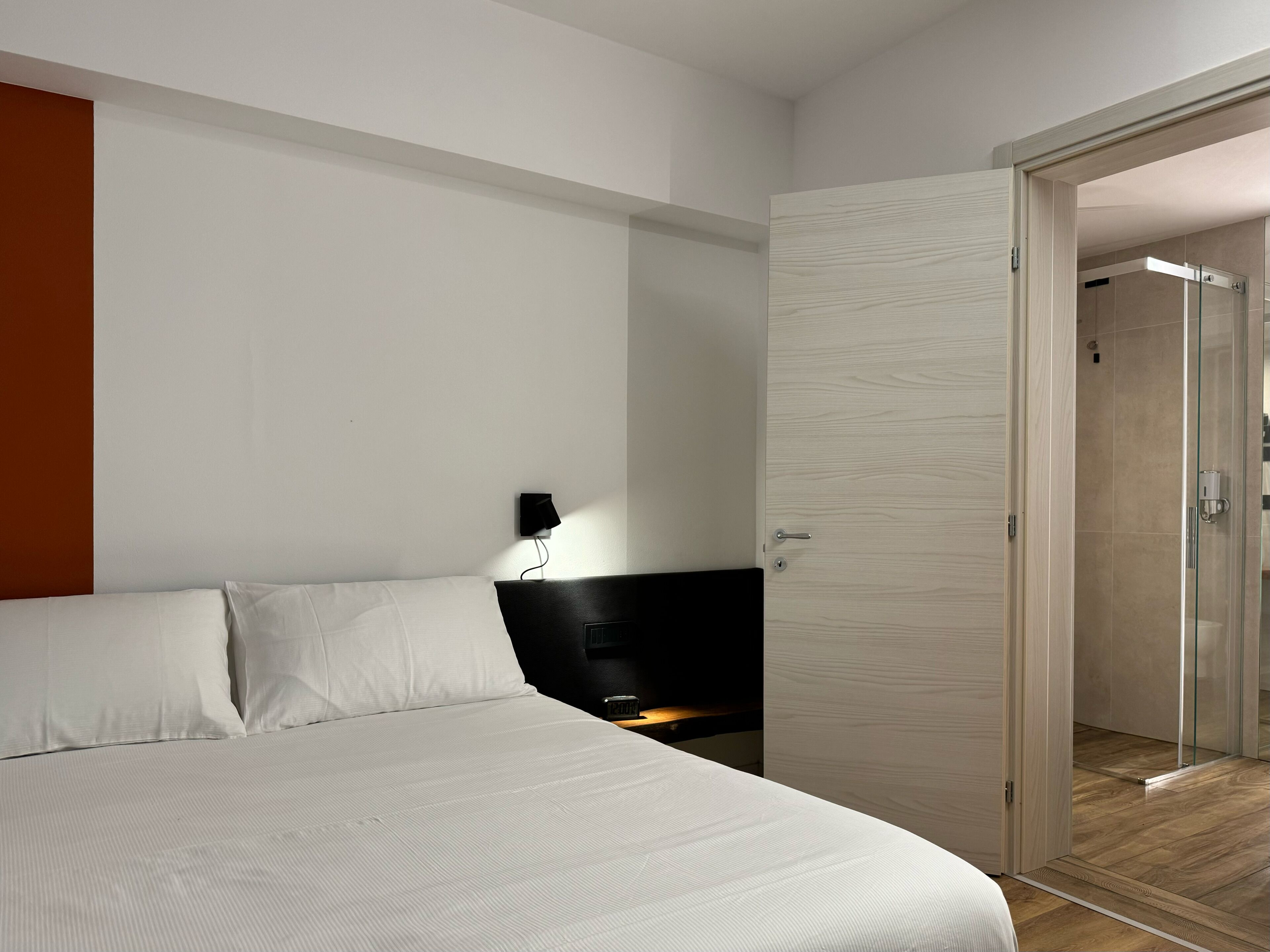 Une chambre d’hôtel moderne avec un grand lit, une table de chevet, une lampe fixée au mur et une porte en verre donnant accès à une cabine de douche.