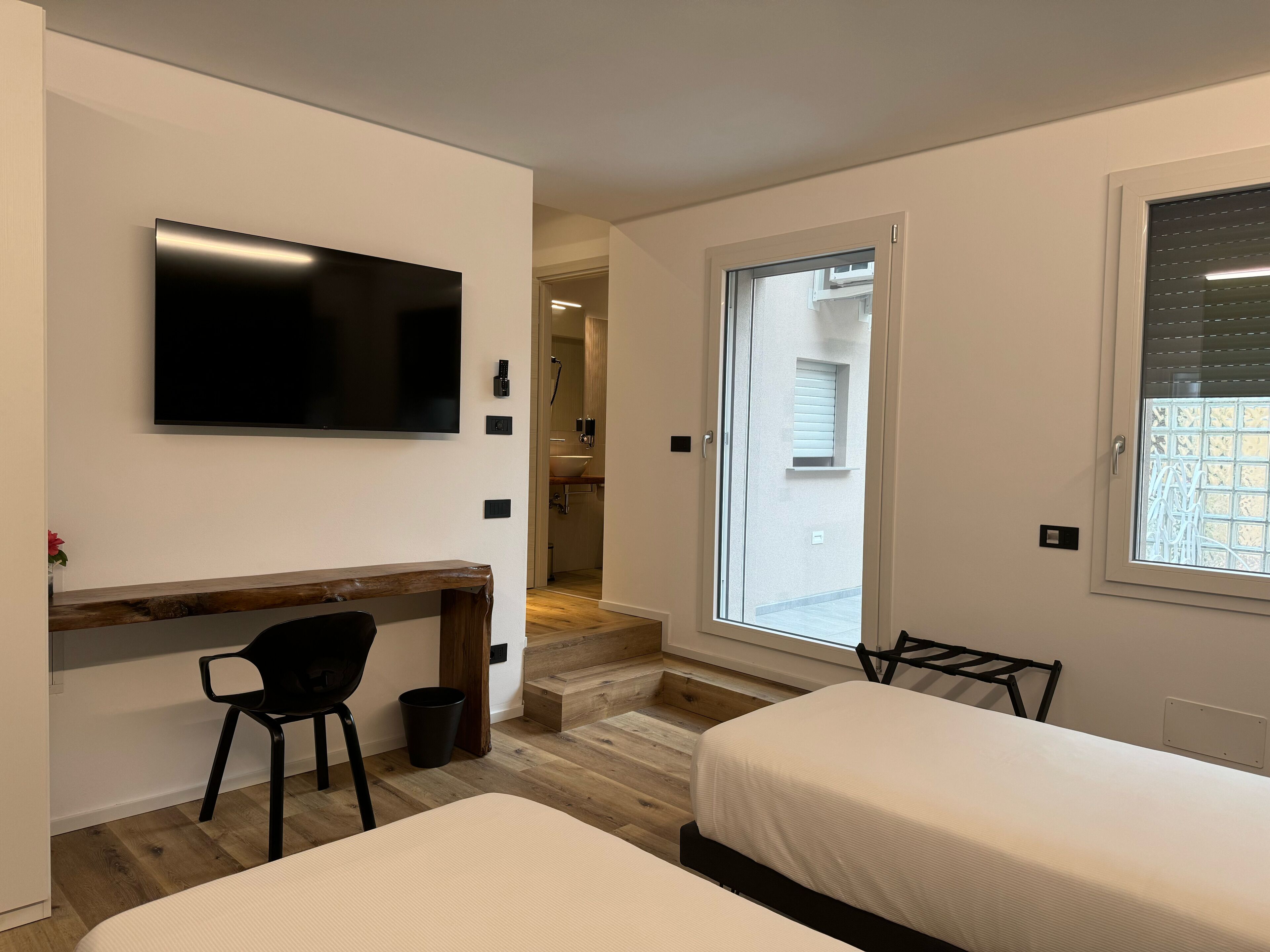 Un dormitorio moderno con televisor, escritorio, dos camas, una silla y un baño visible a través de una puerta abierta.