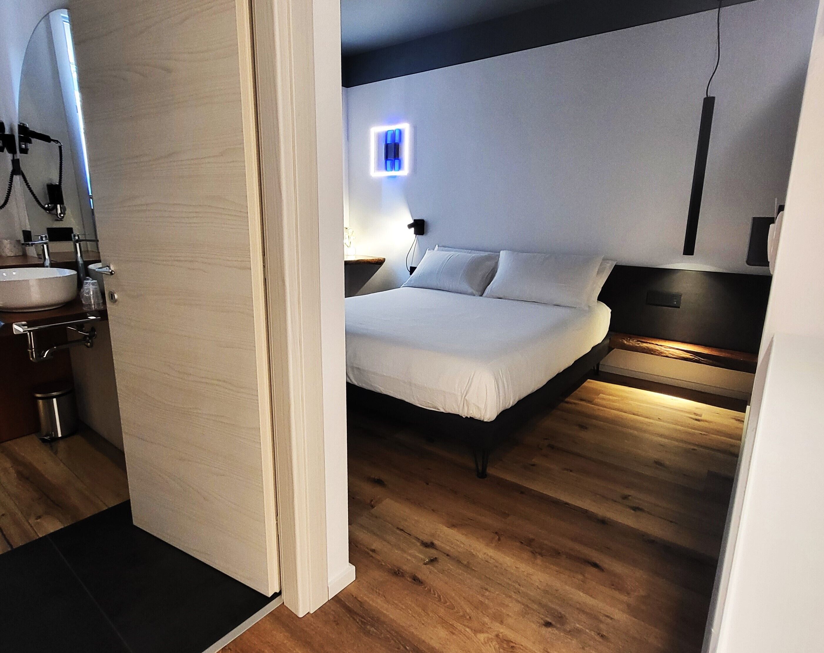 A modern bedroom with a wooden floor, a bed with white bedding, a nightstand, and a wall-mounted light.