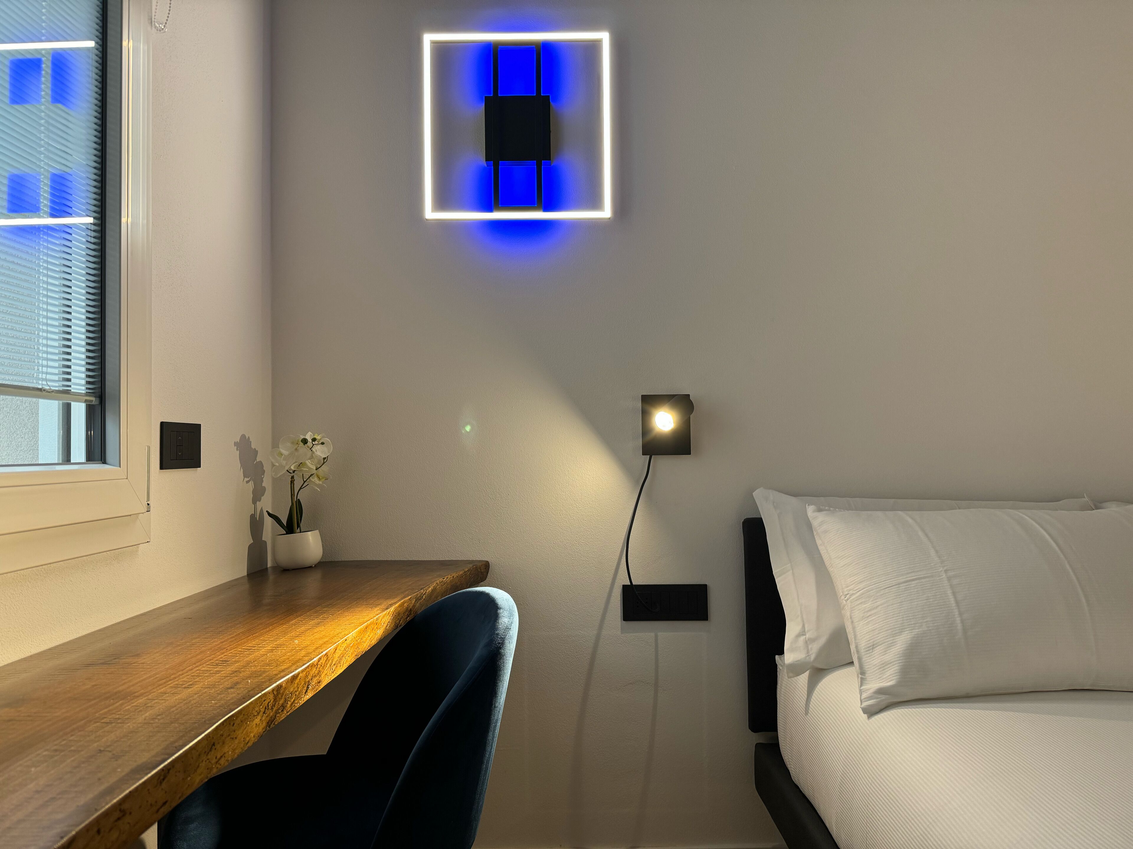 A bedroom with a bed, a desk, and a chair. There is a wall-mounted light fixture above the desk and another on the wall beside the bed.