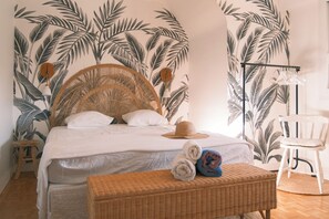 Une chambre avec un lit, une table d’appoint en osier sur laquelle sont posées des serviettes, une chaise et un mur orné d’un motif de feuilles tropicales.