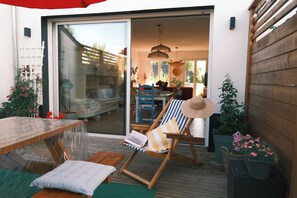 Une terrasse en bois avec une table et des chaises, une porte-fenêtre coulissante donnant sur un espace intérieur et des plantes en pot.