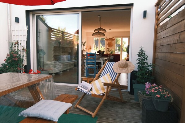 A wooden deck with a table and chairs, a sliding glass door leading to an interior space, and potted plants.