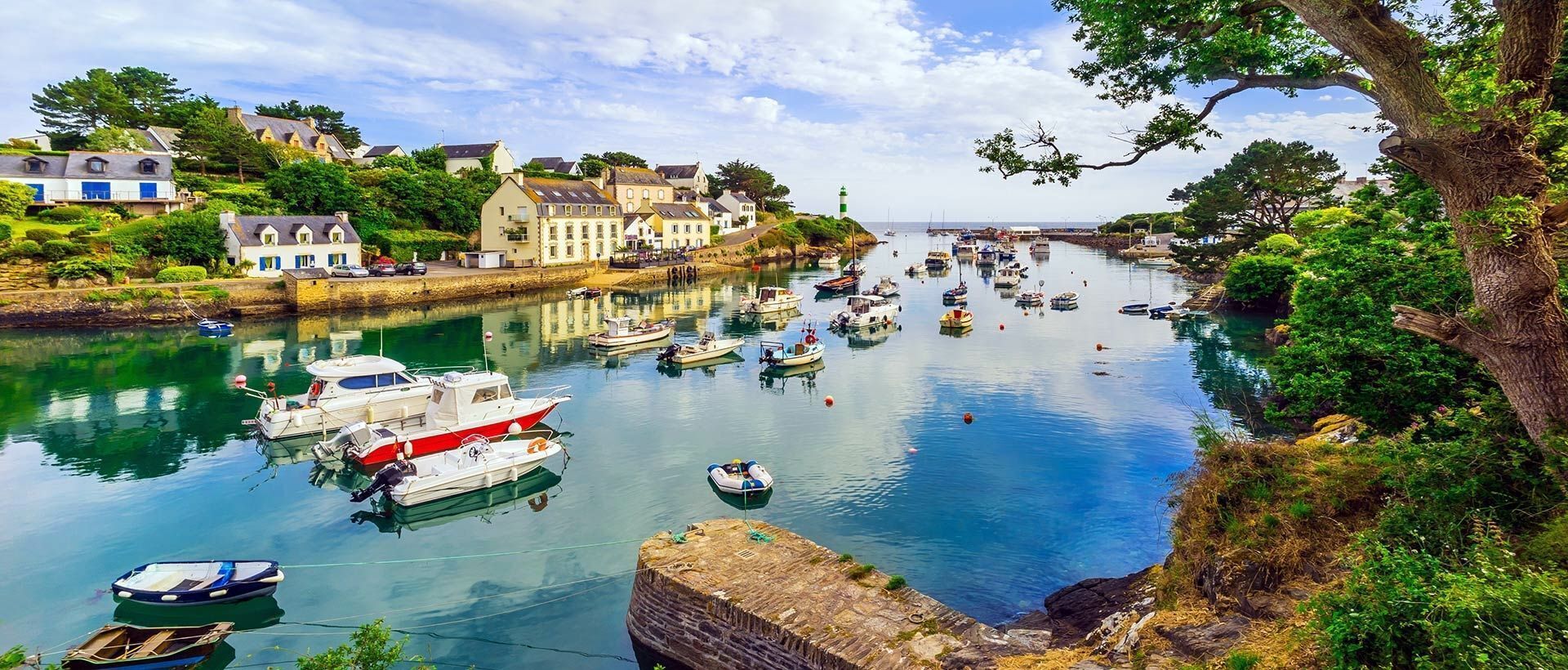 A harbor with boats docked, buildings along the shore, and a lighthouse in the distance.
