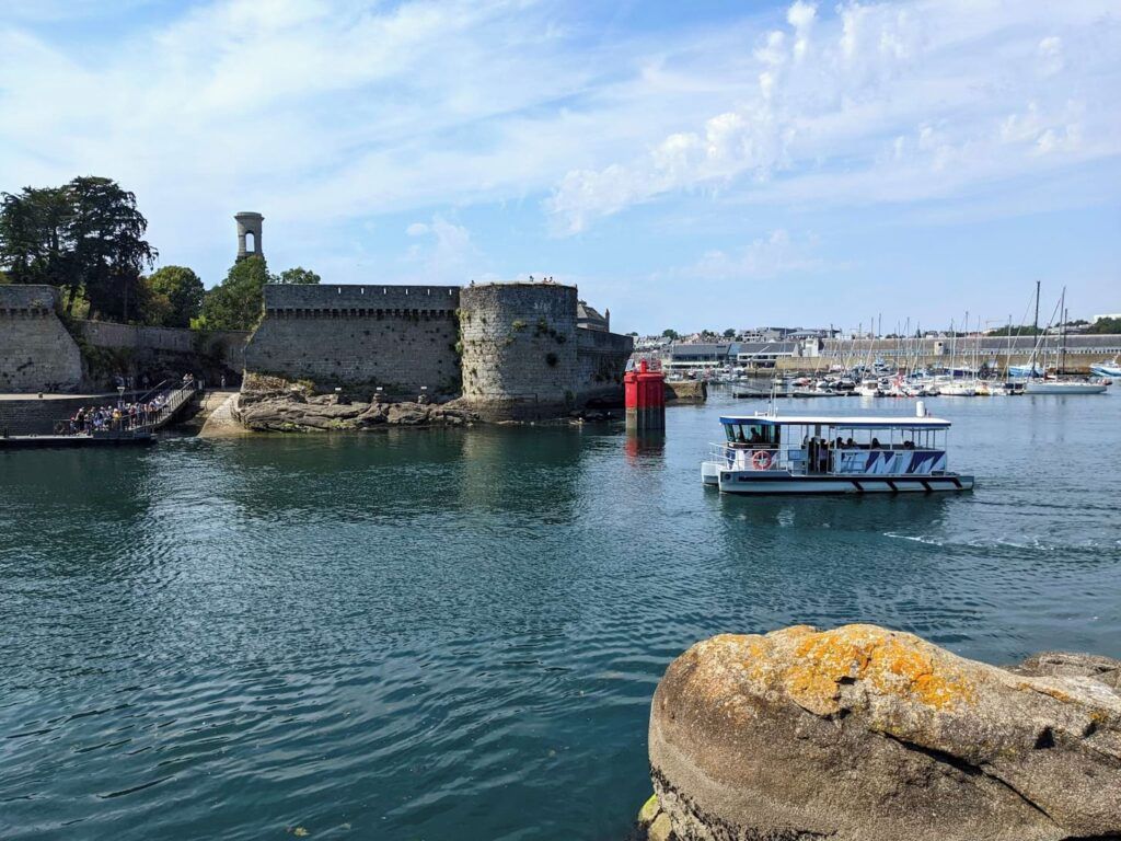 Escena costera con un muro de piedra histórico, un faro y un barco atracado cerca de la orilla.