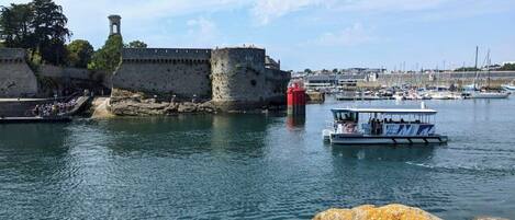 Escena costera con un muro de piedra histórico, un faro y un barco atracado cerca de la orilla.