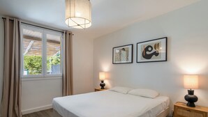 Ein Schlafzimmer mit einem Bett, zwei Nachttischlampen, einem Fenster mit Vorhängen und zwei gerahmten Kunstwerken an der Wand.