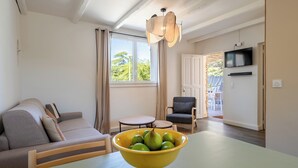 A modern living room with a sofa, a dining table with a yellow bowl of fruit, a grey armchair, and a TV mounted on the wall. - Rental with pool and AC in Provence n°9 (La Roque-sur-Pernes)