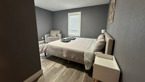 Quarto com cama, mesa de cabeceira, cadeira e janela com persianas.