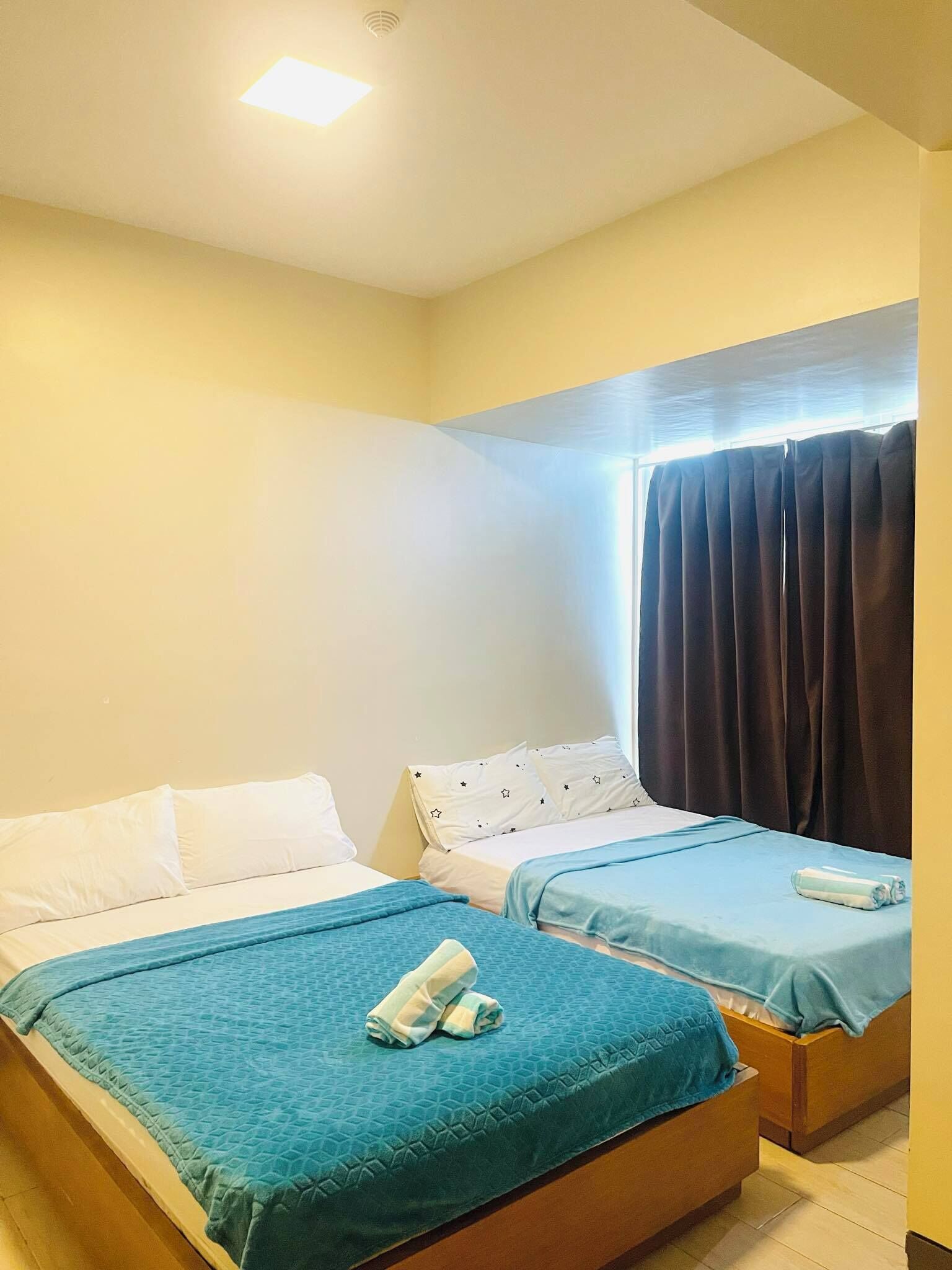 A hotel room with two beds, each with a blue bedspread and white pillows. There is a window with dark curtains, and a ceiling light is visible.
