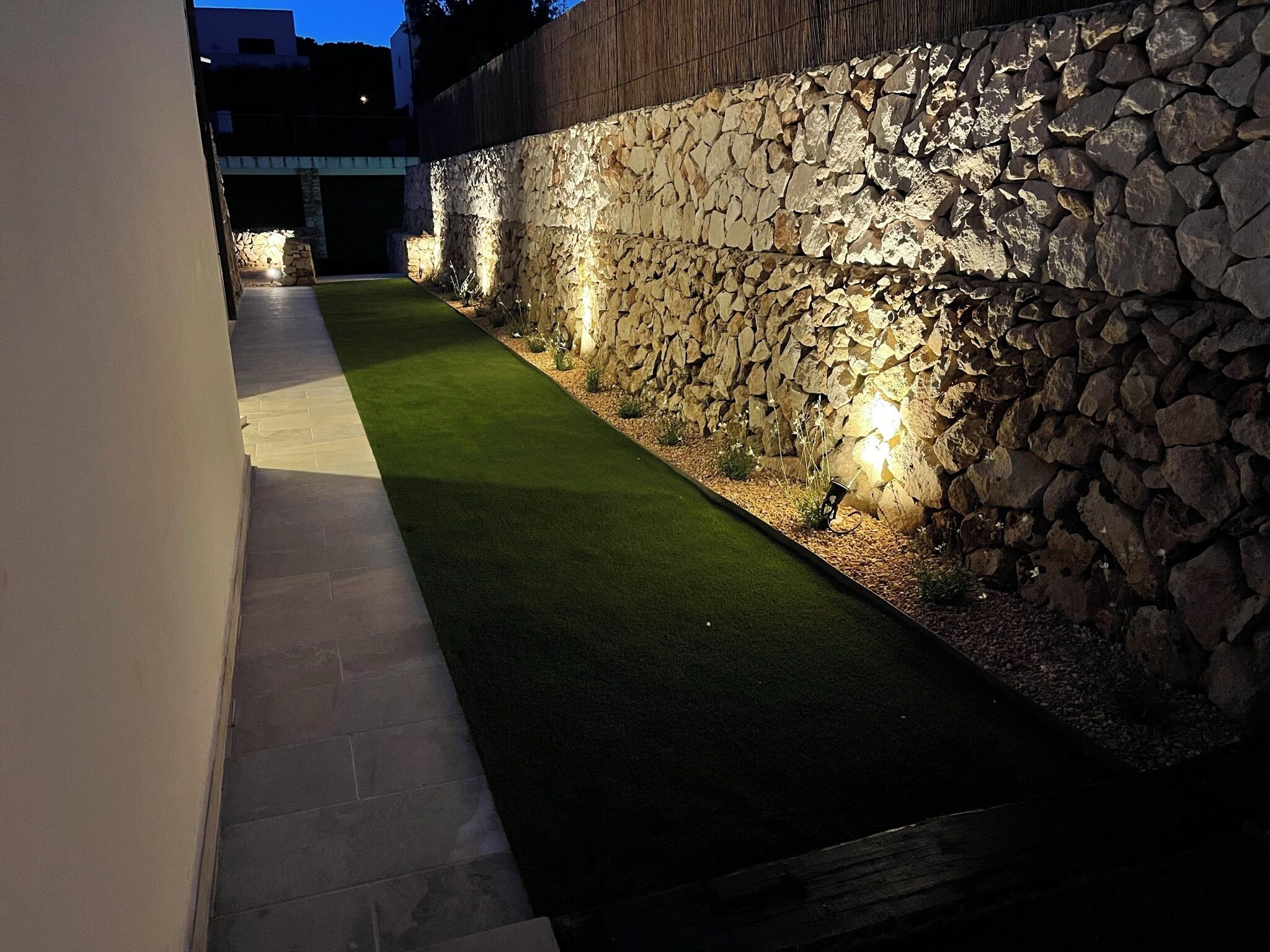 A well-lit outdoor area with a stone wall, a paved walkway, and a grassy section.