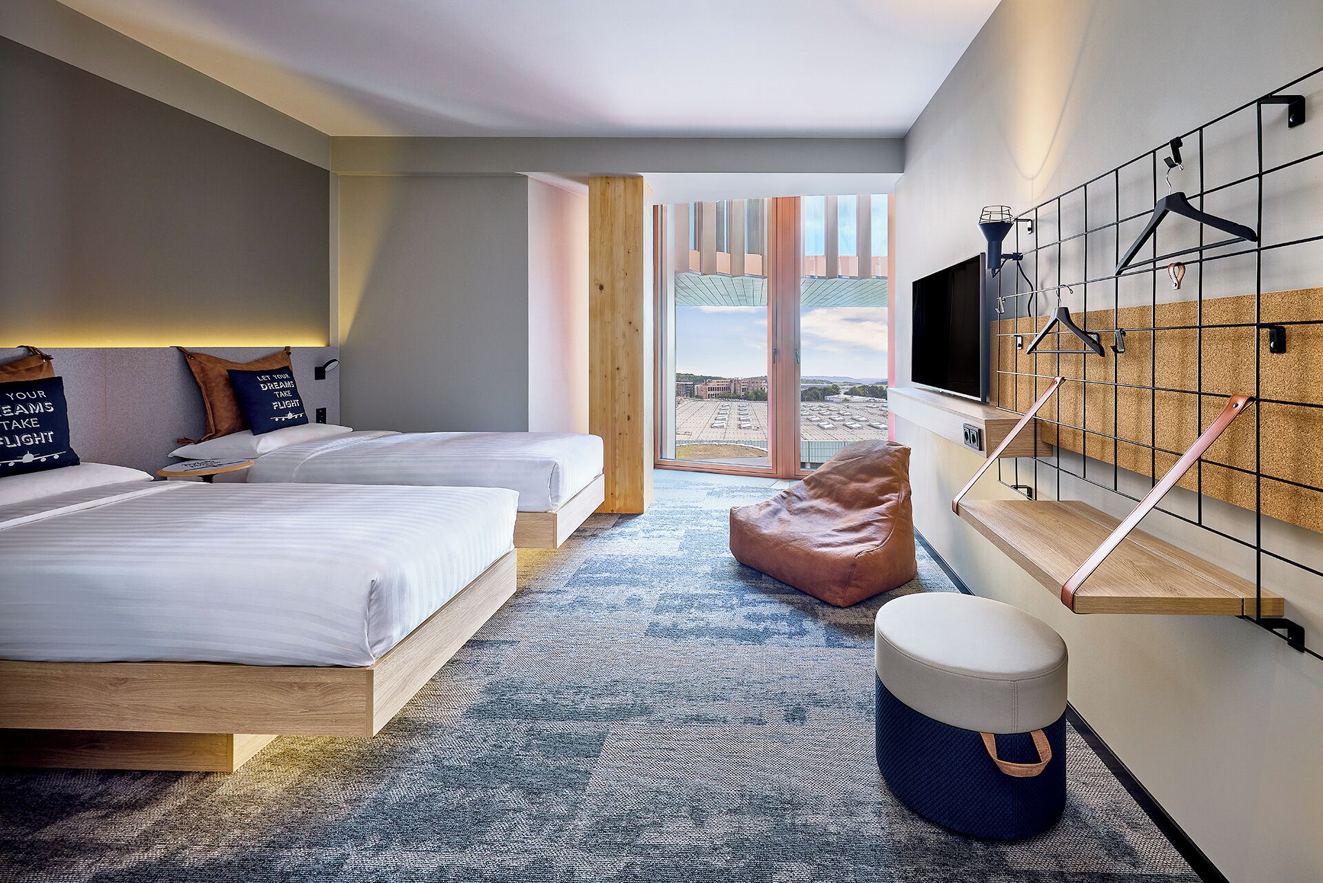 A modern hotel room with two beds, a large window, a TV, and a wall-mounted rack.